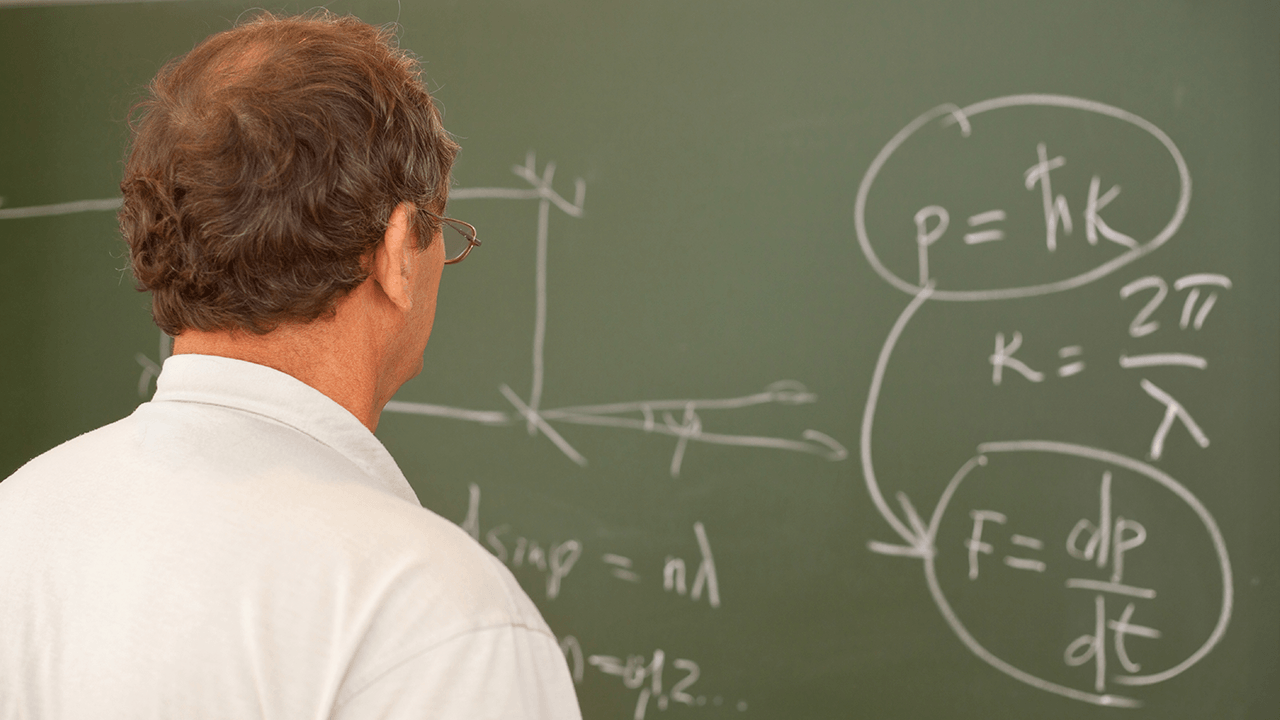 A professor studies equations on a chalkboard covered with mathematical formulas.