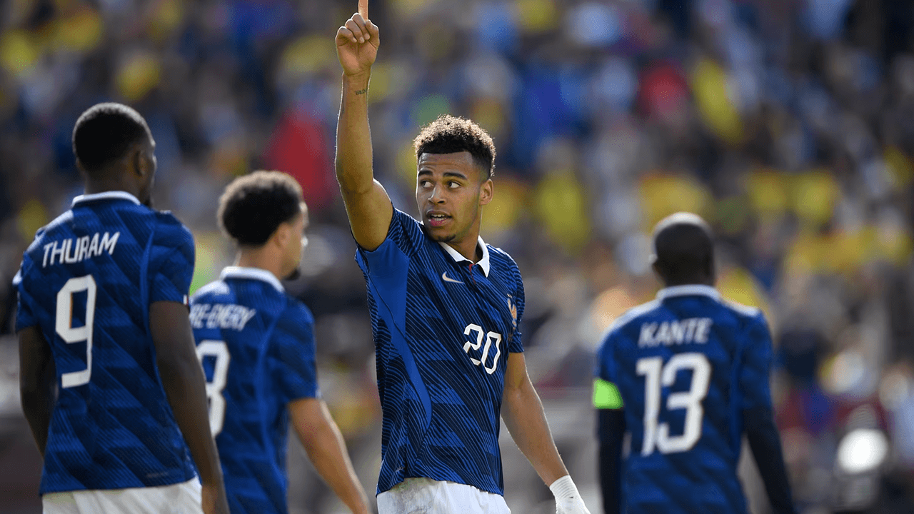 Kylian Mbappé of the French national team raises his right index finger skyward in celebration during a soccer match, with teammates Thuram (No. 9) and Kanté (No. 13) visible against a blurred stadium crowd.