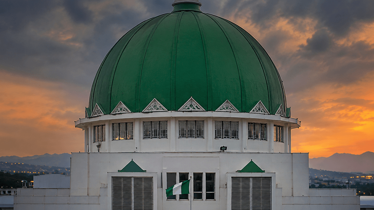 Close-up view of the Nigerian National Assembly building, showcasing its prominent green dome and the Nigerian flag flying in front against a dramatic sunset sky.