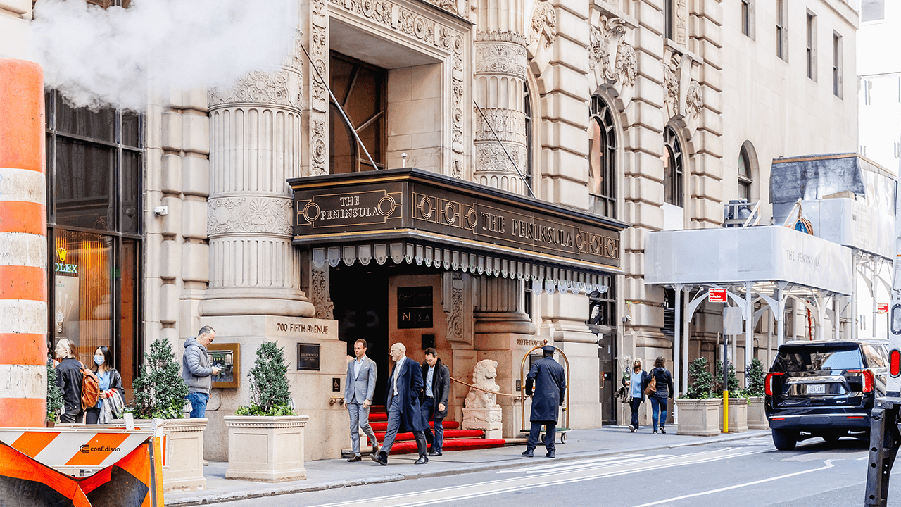 The entrance of The Peninsula hotel on Fifth Avenue in Manhattan is shown with pedestrians, hotel staff, and light street activity outside the historic building.